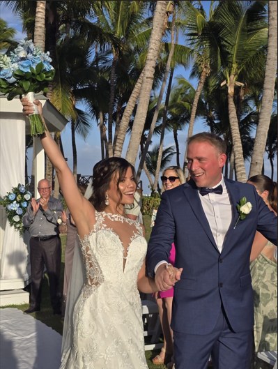 Bride raising her bouquet as the couple walks back down the aisle under palm trees
